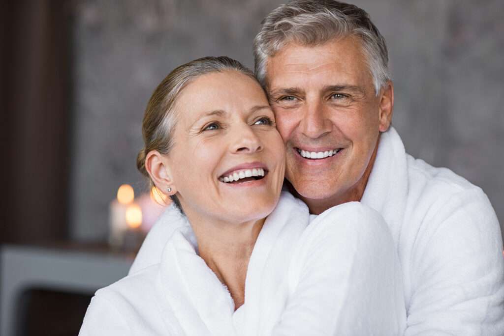 Smiling husband embracing cheerful wife from behind at spa. Laughing mature couple enjoying a romantic hug at wellness center after massage. Senior man and woman in white in bathrobe relaxing at spa.